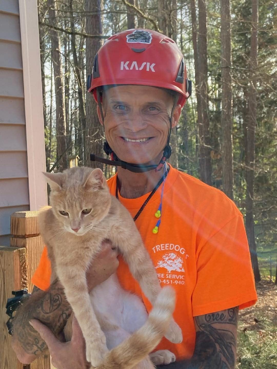 Man in orange safety shirt and red helmet holding a tan cat outdoors by a wooded cabin