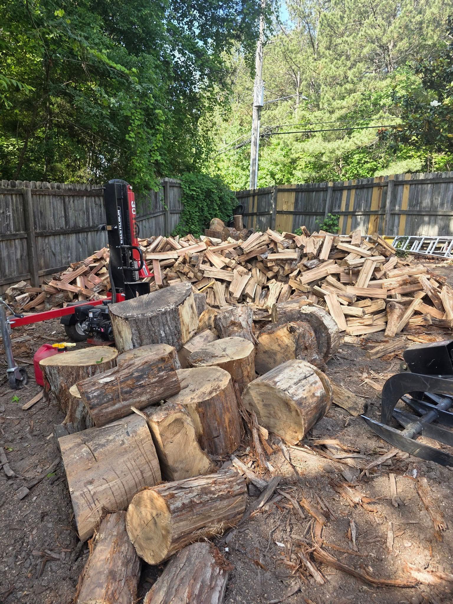 Woodpile of split logs beside a red log splitter in a fenced backyard, with trees in the background