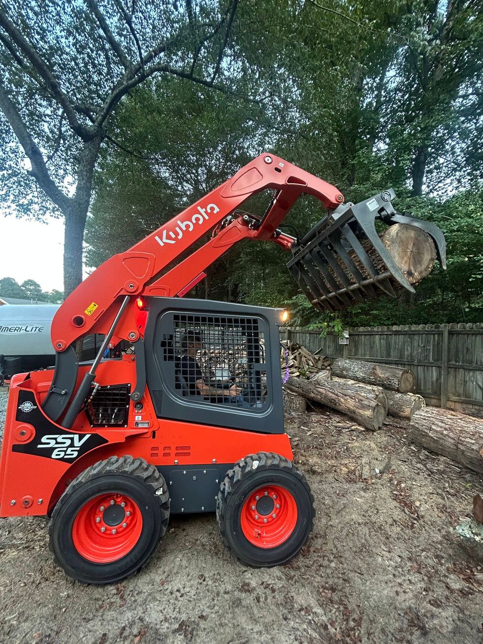 Red compact loader lifting a bucket of logs beside a woodpile under trees