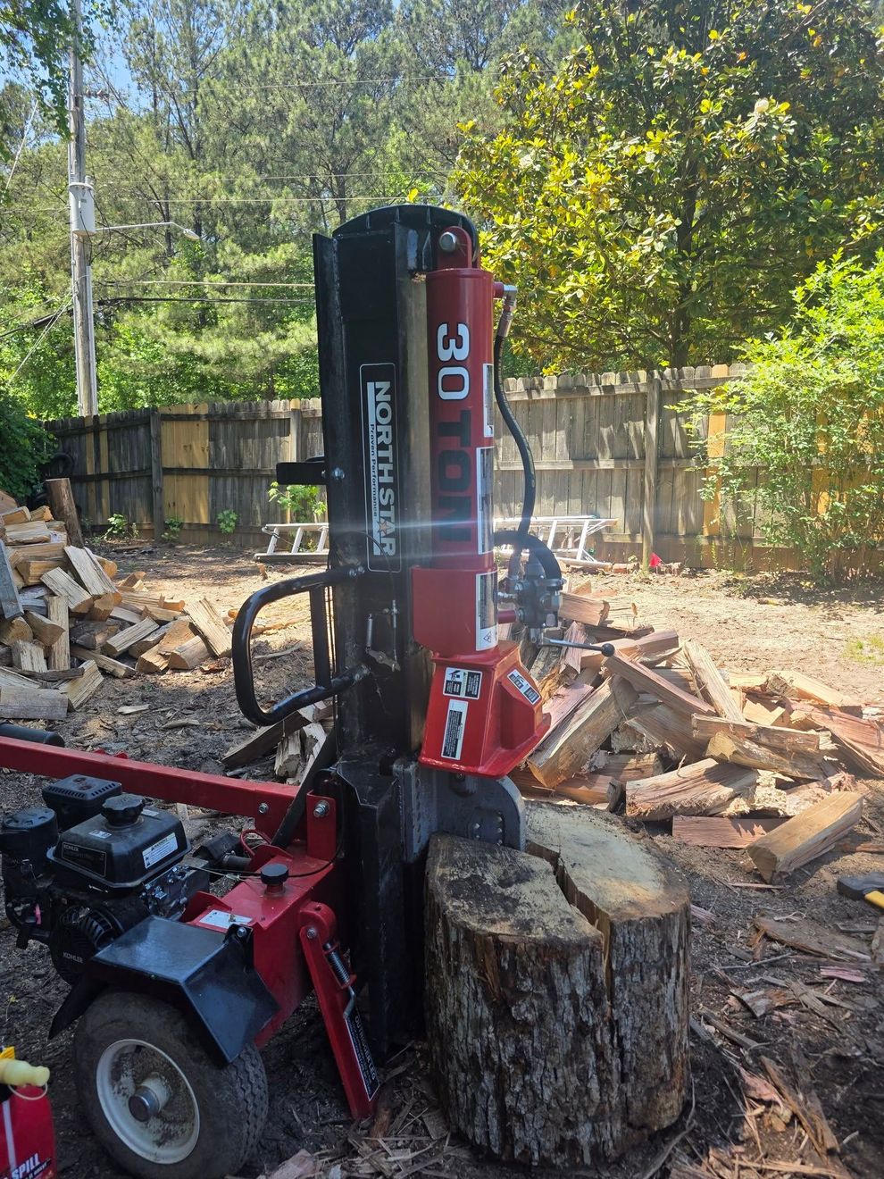 Red wood splitter lifting a log in a sunny backyard with stacked cut wood and trees nearby