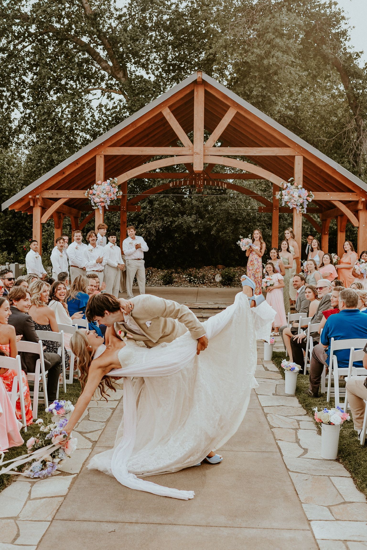 Groom dips bride, kissing, on outdoor wedding aisle, before guests and wooden gazebo.