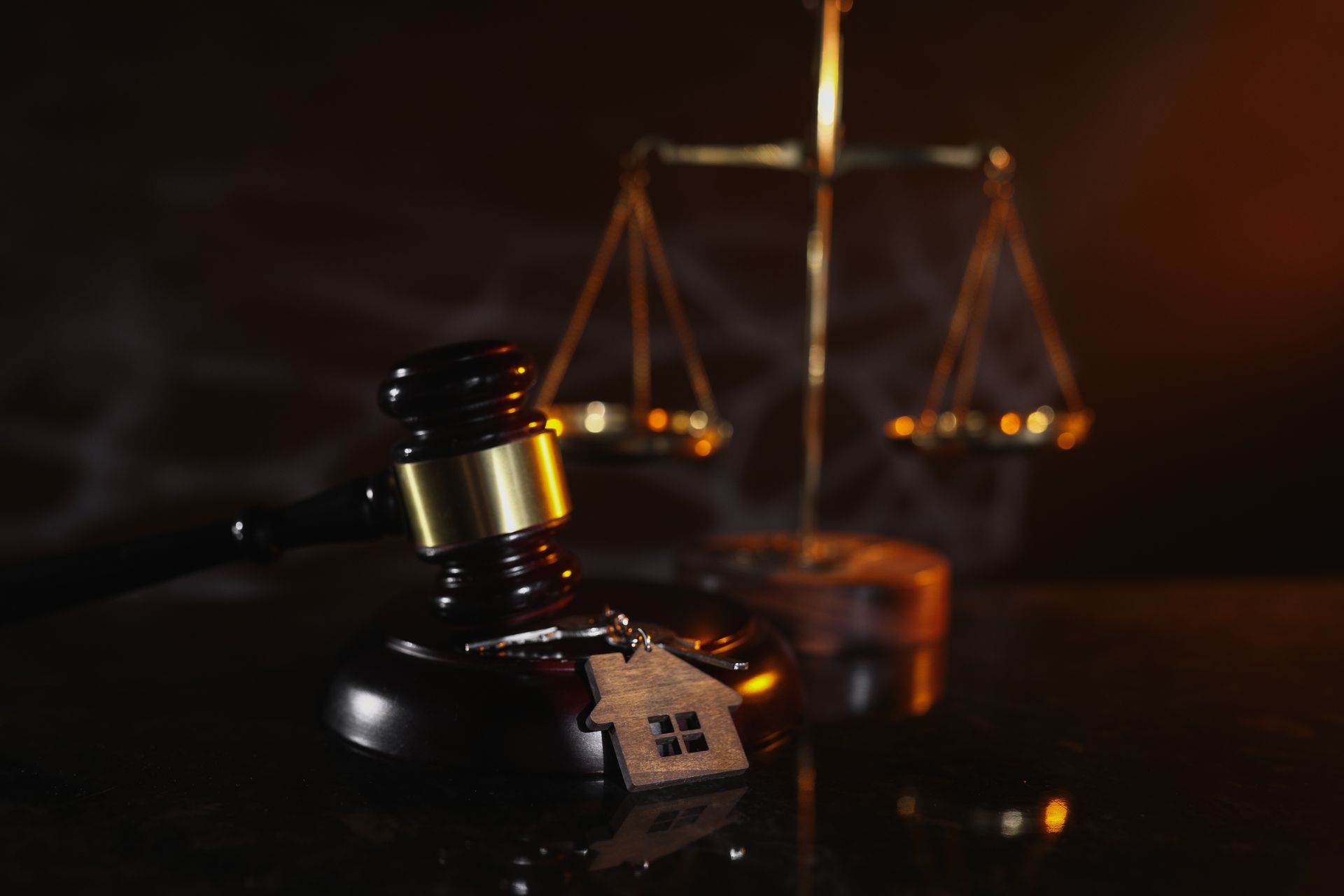 Gavel and miniature house rest next to the scales of justice in a dimly lit setting.