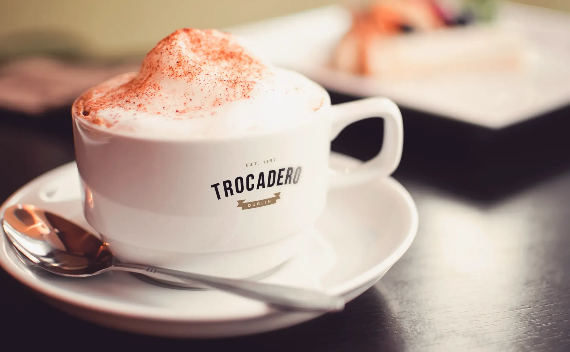 Cup of cappuccino with foam, dusted with spice, on saucer, spoon, and dark table. 