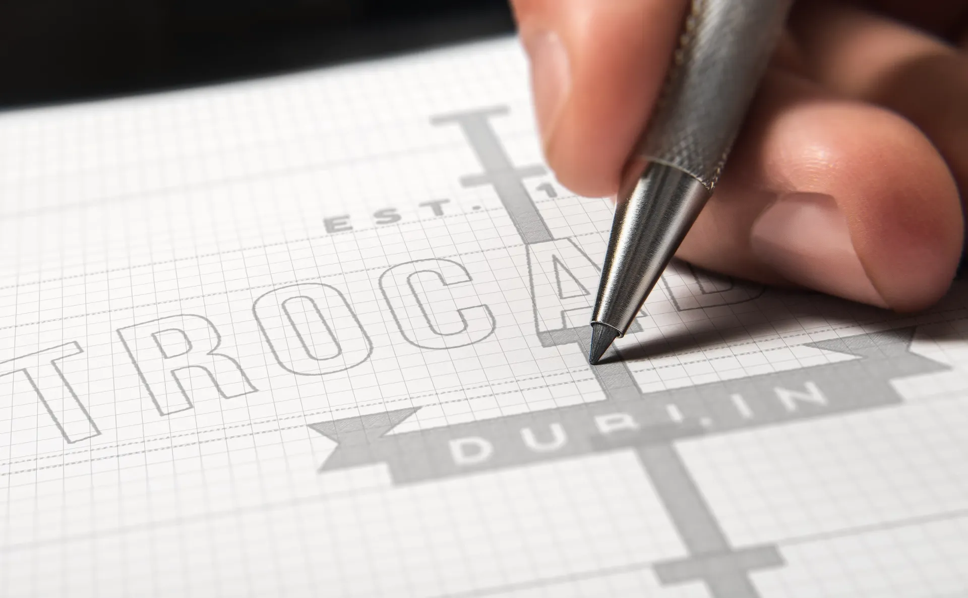 Hand writing with a pencil onto a grid paper layout, spelling the word 