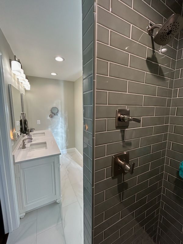 A bathroom featuring a shower with dark gray subway tiles and a vanity with a white countertop and dual sinks.