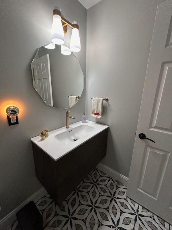 A modern powder room featuring a wall-mounted dark vanity, white countertop, gold faucet, geometric floor, and round mirror.