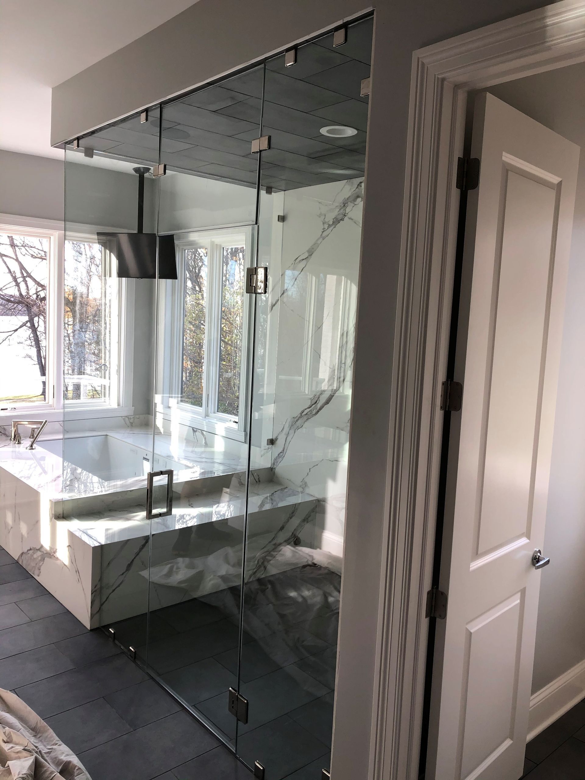 Glass shower enclosure in a bathroom, next to a bathtub. White marble and white door.