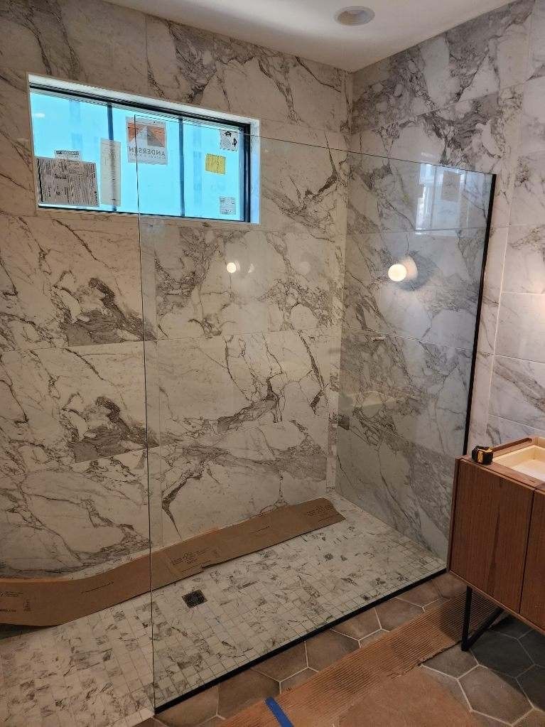 Modern bathroom with marble tile walls, glass shower, window, and wood vanity.