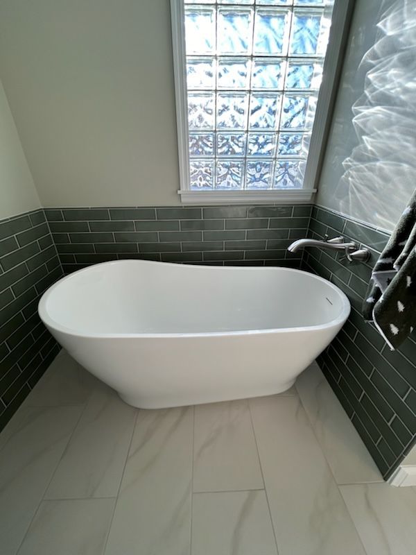 A white freestanding bathtub set against dark green subway tile walls in a bathroom with light tile flooring.