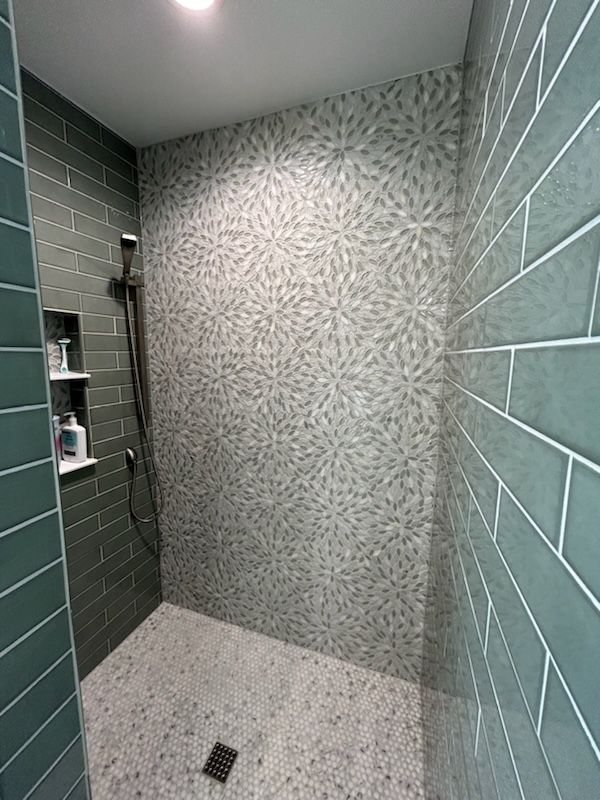 A shower with teal subway tiles, a mosaic-patterned back wall, and small stone floor tiles, featuring a recessed shelf.