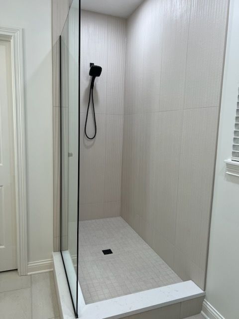 A walk-in shower with light-colored tile walls, a light stone floor, and a black showerhead and drain.