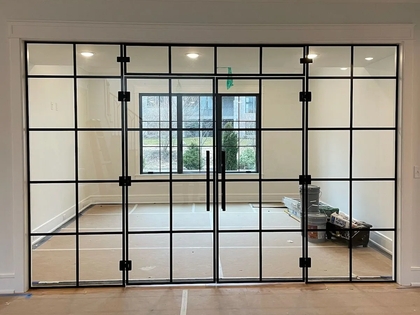 Glass-paneled doors with black frames open to a room with a window, white walls, and a paper-covered floor.