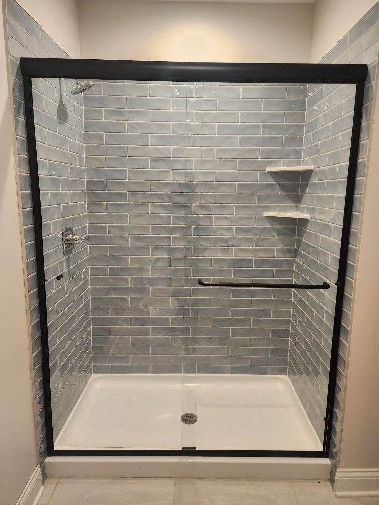 A modern shower with gray brick-like tile, glass doors, black frame, and white shower pan.