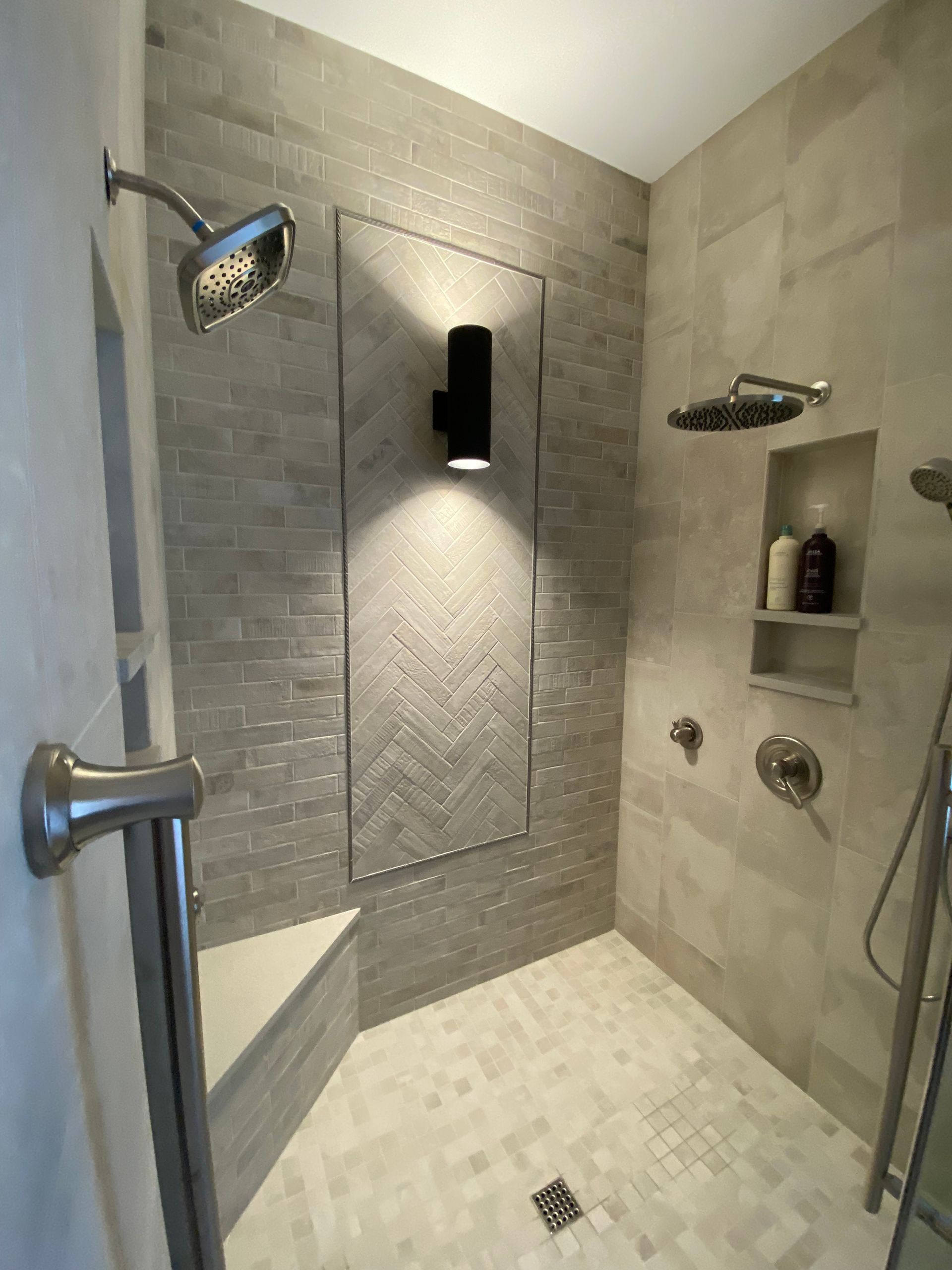Accessible shower with gray tile, bench, and grab bar. Includes multiple shower heads, and recessed storage.