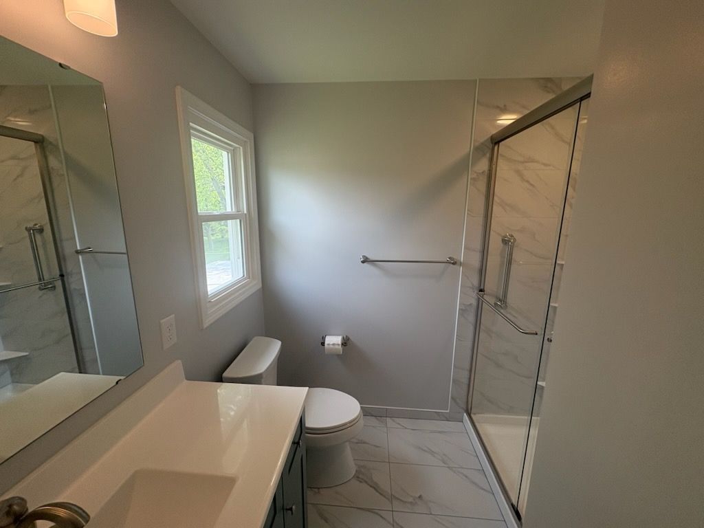 Bathroom with white vanity, toilet, and marble-look shower. Gray walls and window.
