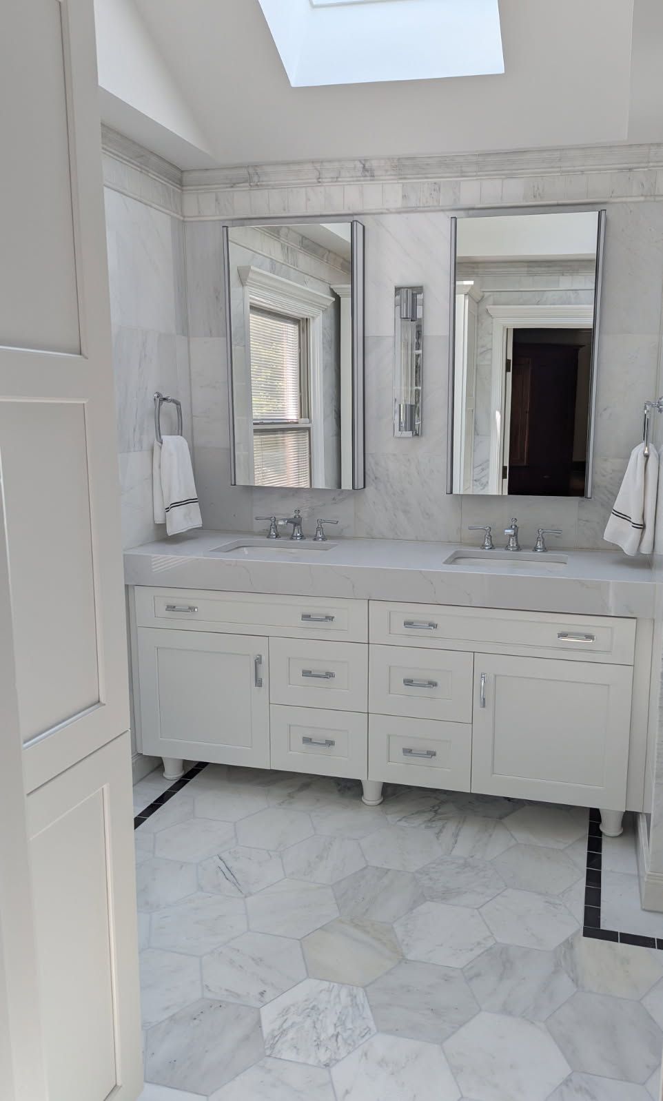 White bathroom with double sinks, mirrors, and marble tile. Skylight above.