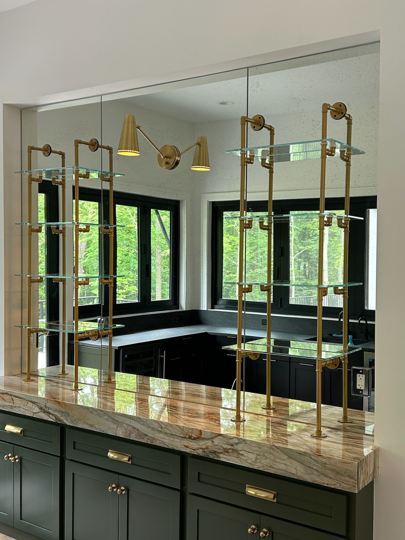 Brass shelving unit with glass shelves, mounted over a dark countertop. A bright kitchen is in the background.