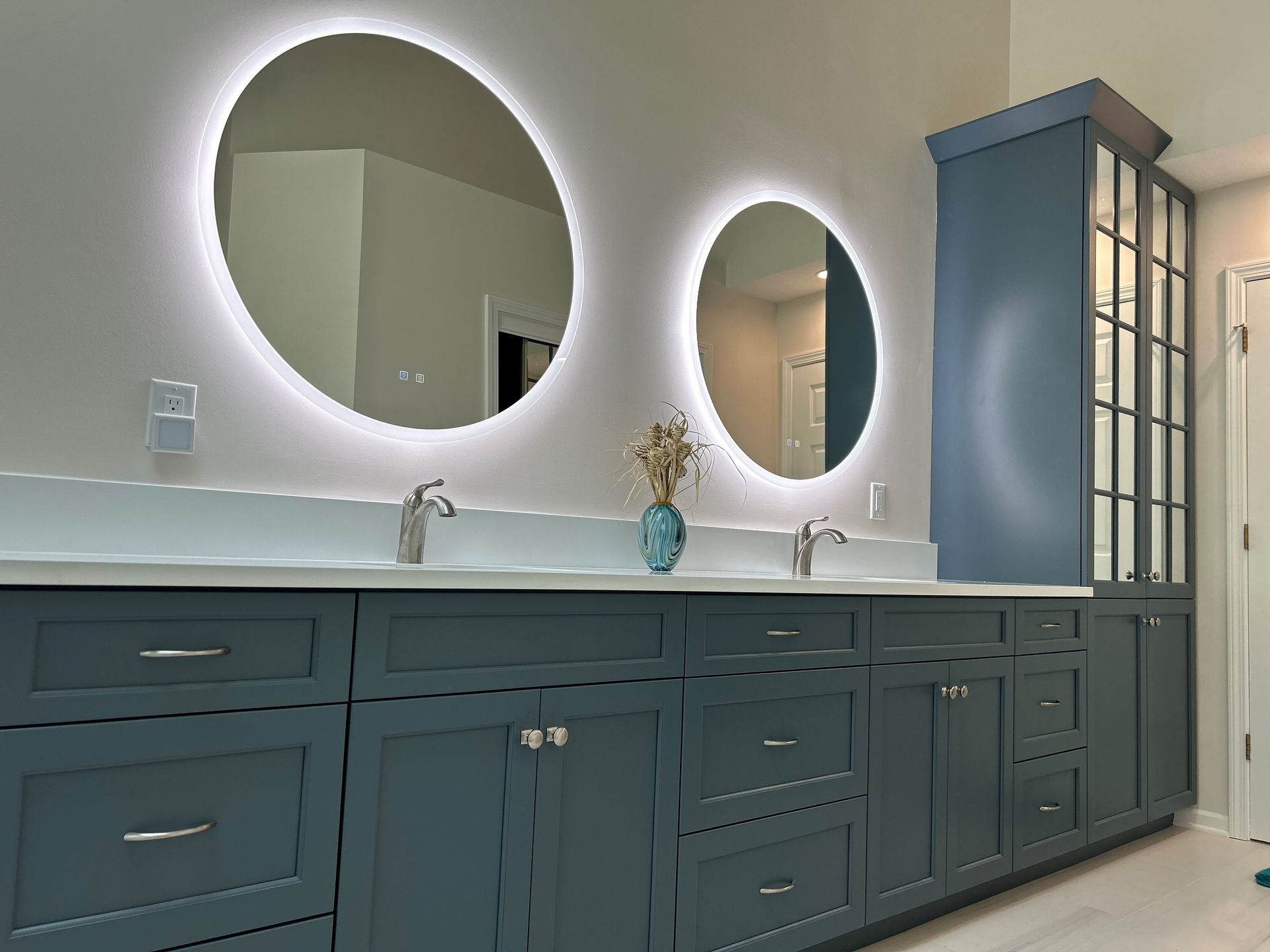 Bathroom vanity with blue cabinets, two round lighted mirrors, and a tall storage cabinet.