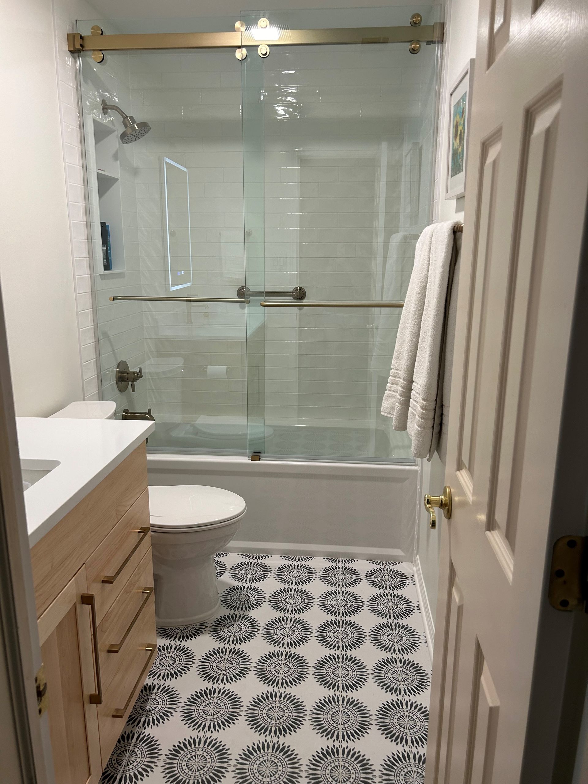 Bathroom with white walls, patterned floor, glass shower, light wood vanity, and a towel on a rack.