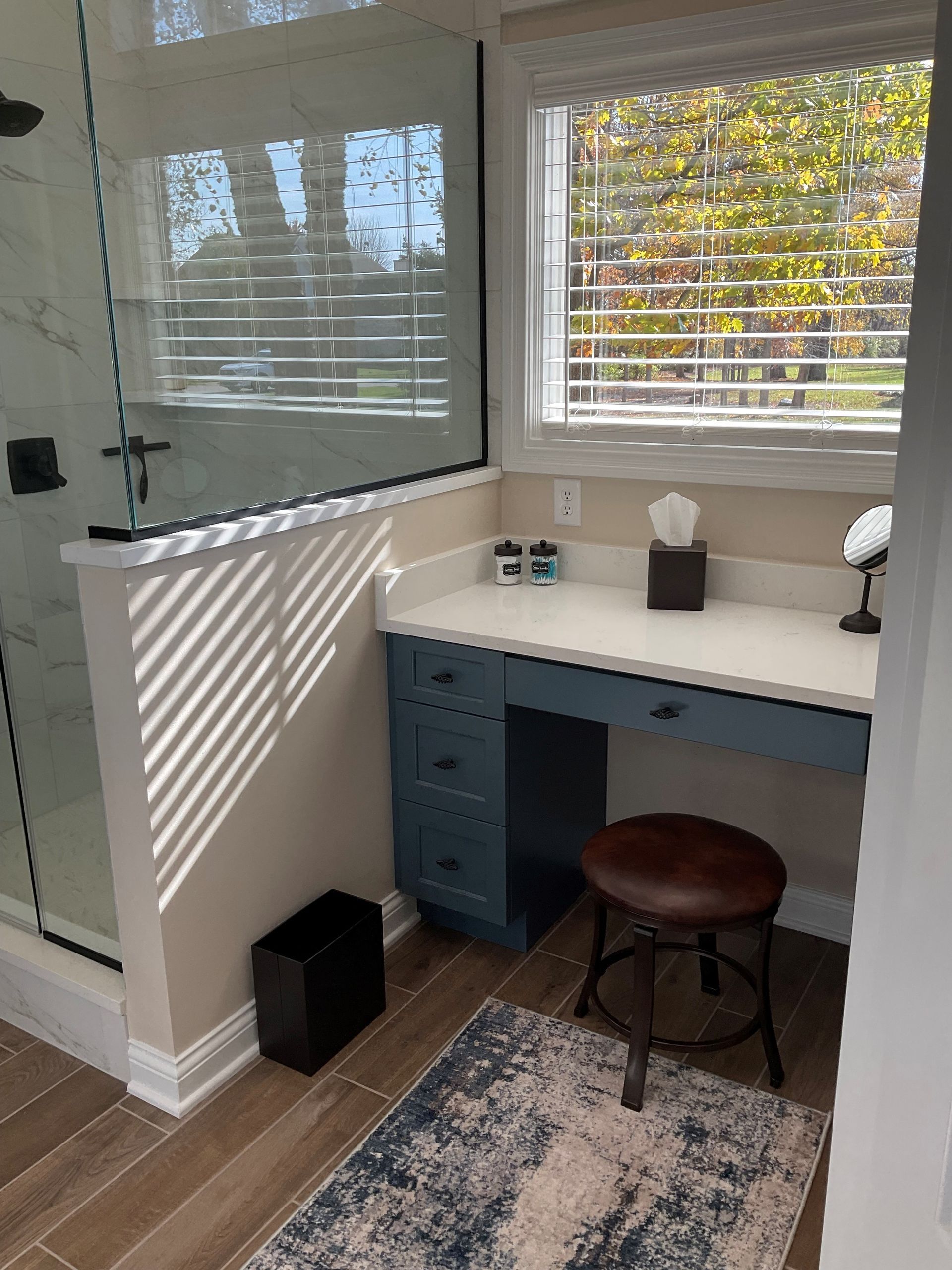 Bathroom vanity with blue cabinets, a small rug, a stool, and a shower with glass doors.