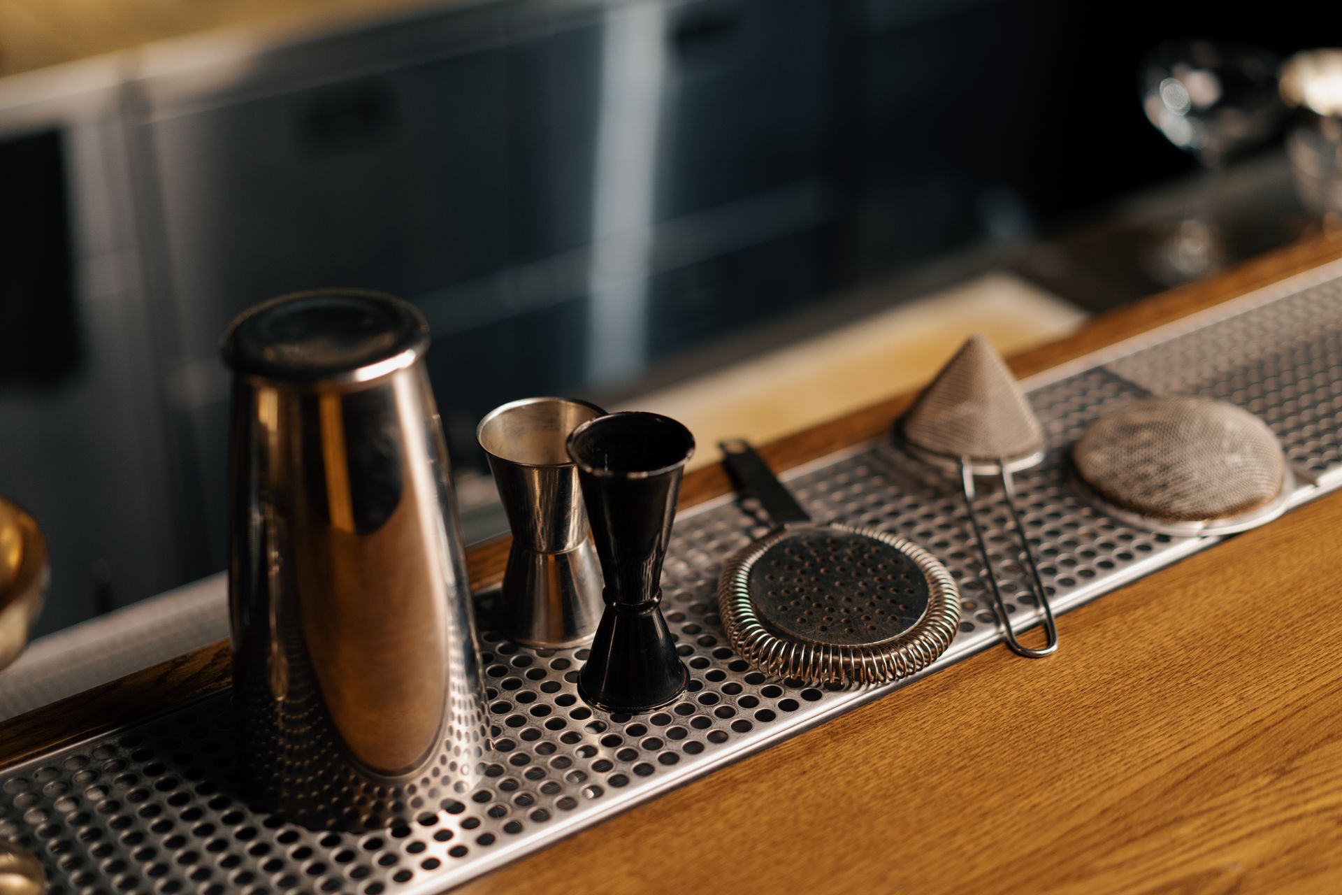 Cocktail bar tools on a metal counter, including shaker, jigger, strainer, and ice scoop