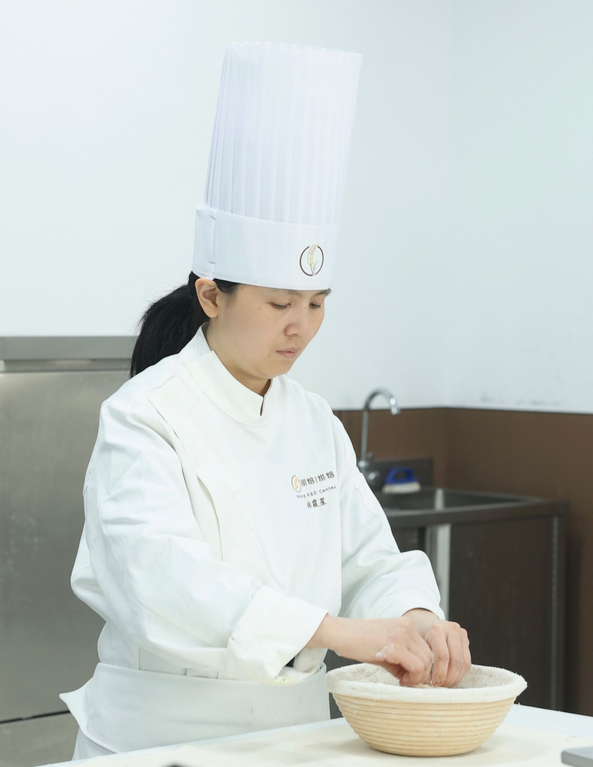 Chef in a white uniform shaping dough in a basket, bright kitchen setting.