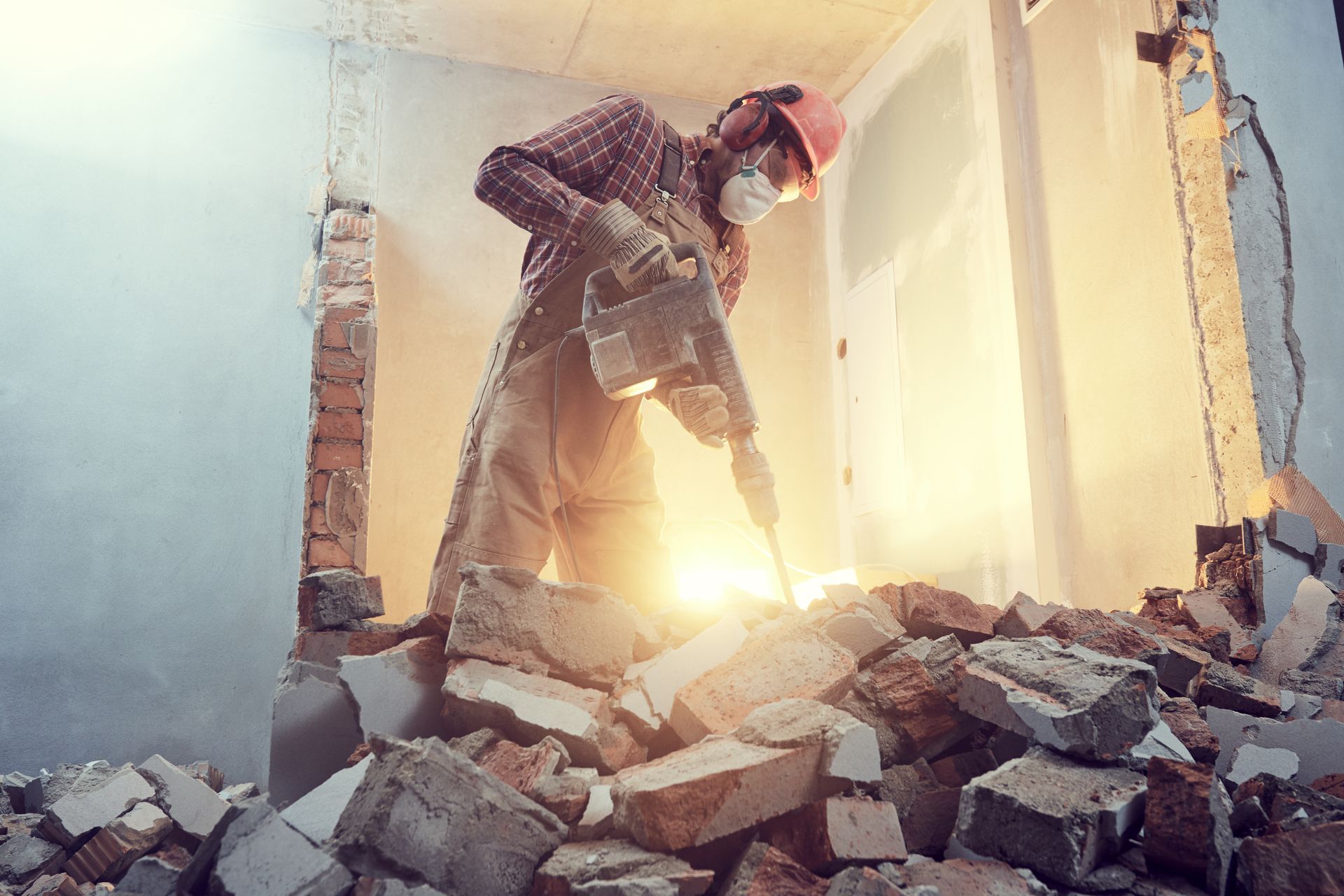 Worker using a jackhammer breaks concrete in a dusty construction site, with bright light behind him