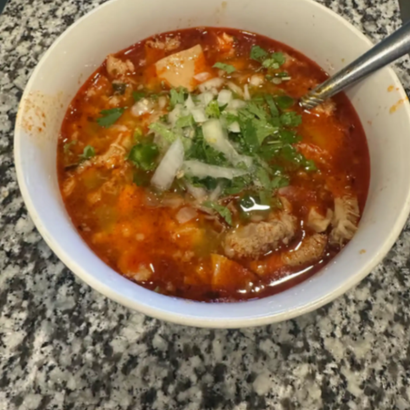 Bowl of red soup topped with chopped onions and cilantro, with a spoon on a granite countertop