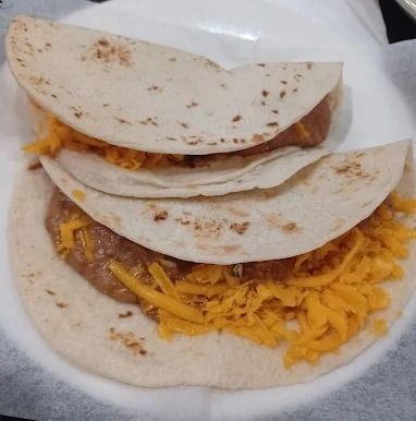 Two folded tacos on white paper, topped with shredded yellow cheese and meat.