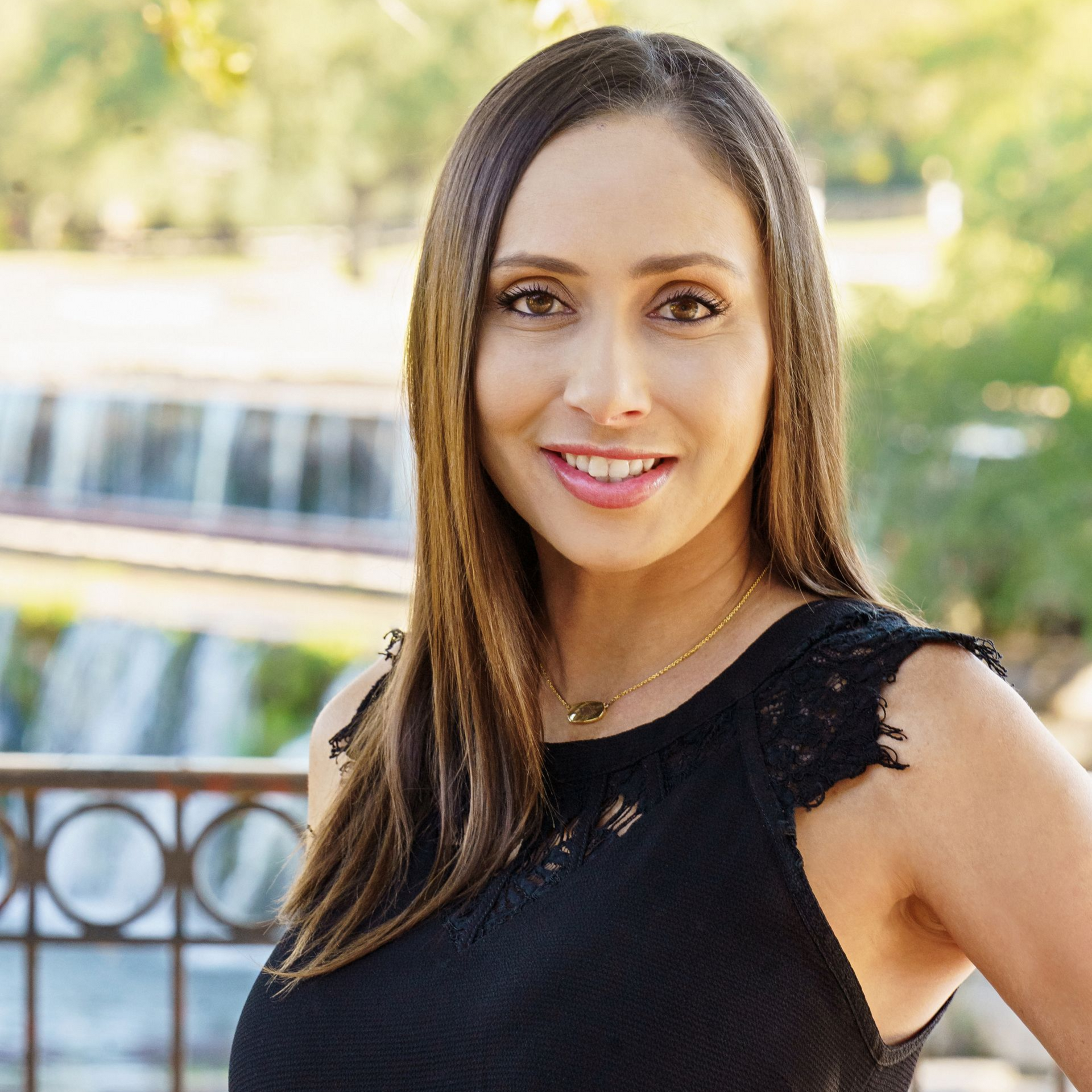 Woman with long brown hair wearing a black top smiles outdoors, with trees and a waterfall in the background.