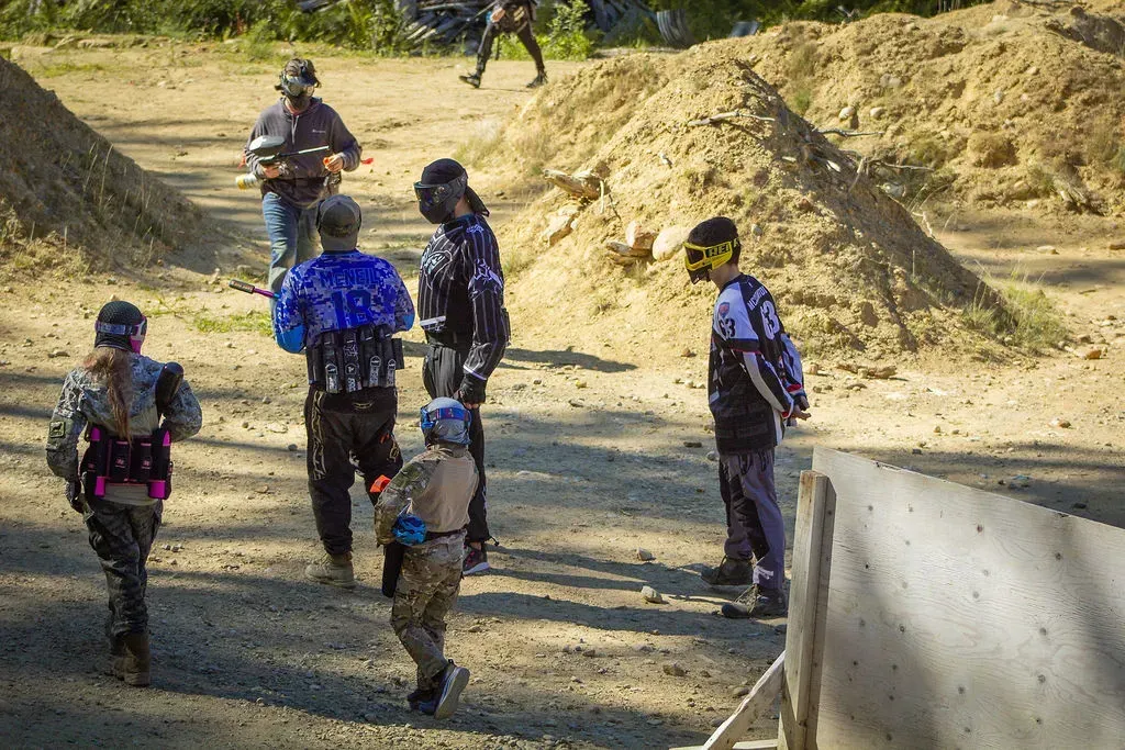 Group of people in paintball gear in a wooded area pose near a decorated totem pole.