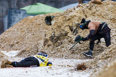 A person playing paintball aims a gun from behind a triangular wooden structure in a forest.