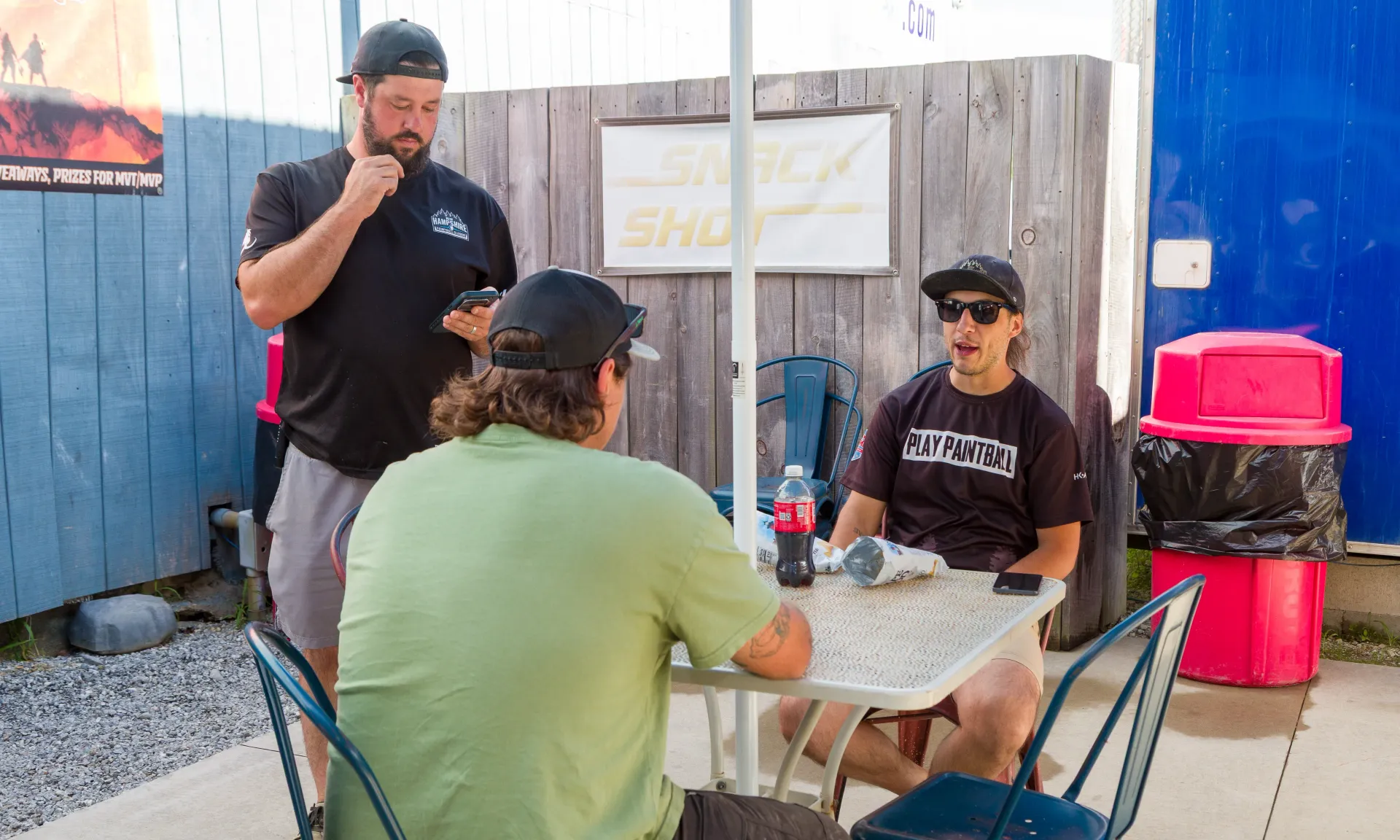 Three people sitting at a table outdoors. One is pondering, another is talking, the third is seated.