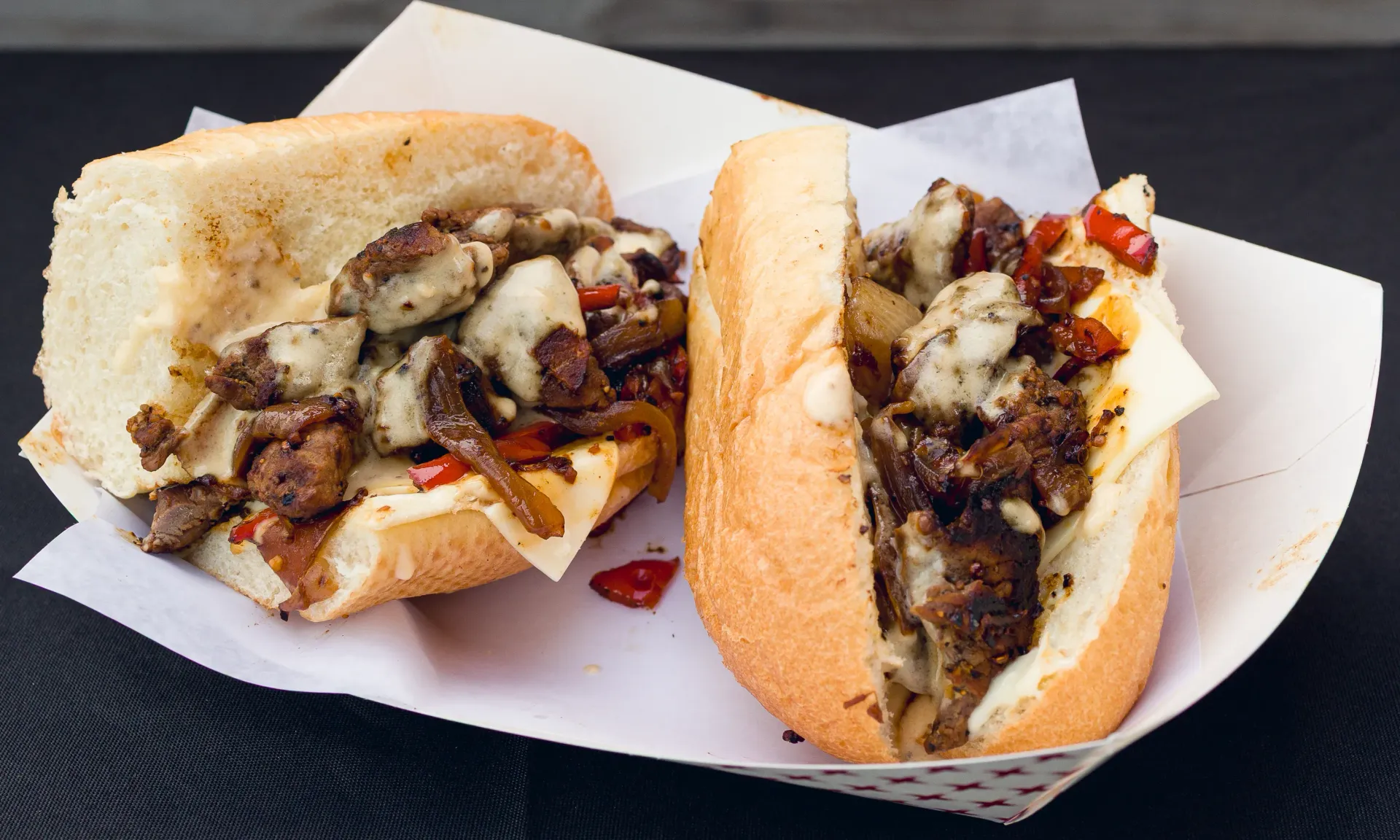 Two halves of a cheesesteak sandwich on a paper-lined tray, with meat, cheese, and peppers visible.