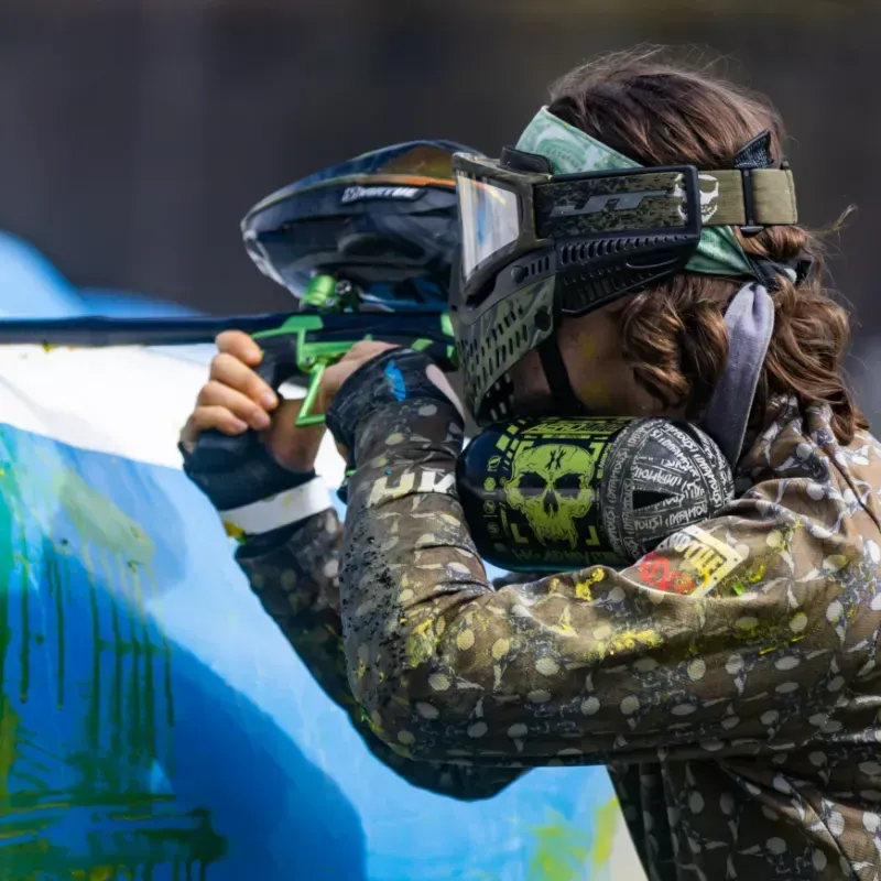 Paintball player aiming green marker, wearing camouflage, mask and holding a skull air tank.