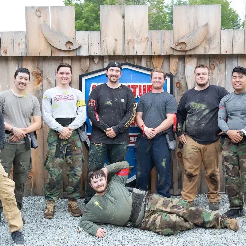 A group of paintball players pose for a photo in front of a wooden structure; one man is lying down.