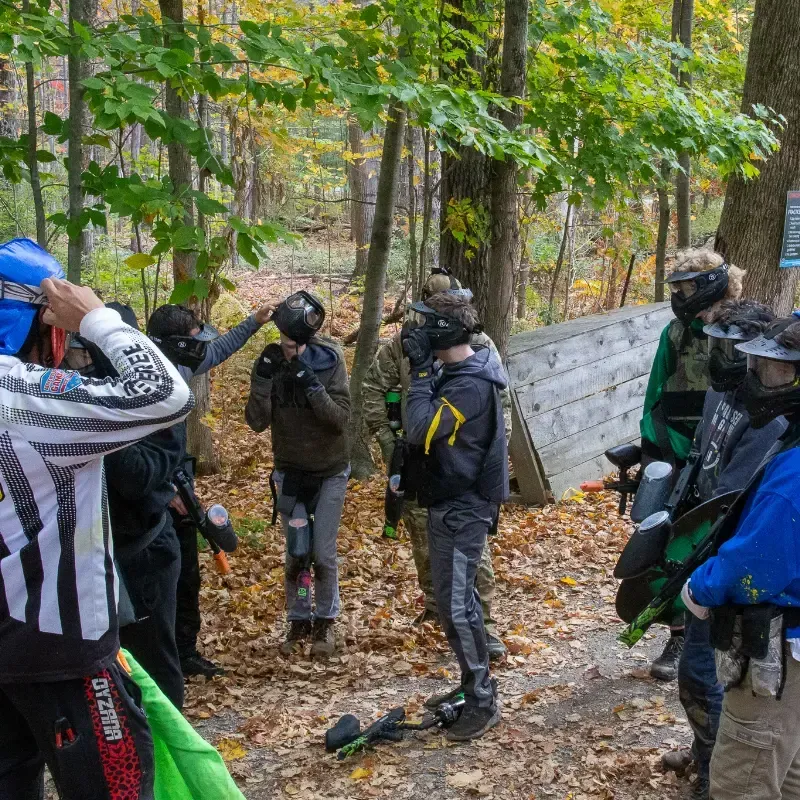 People in paintball gear in a wooded area, some with masks on, holding guns, preparing to play.