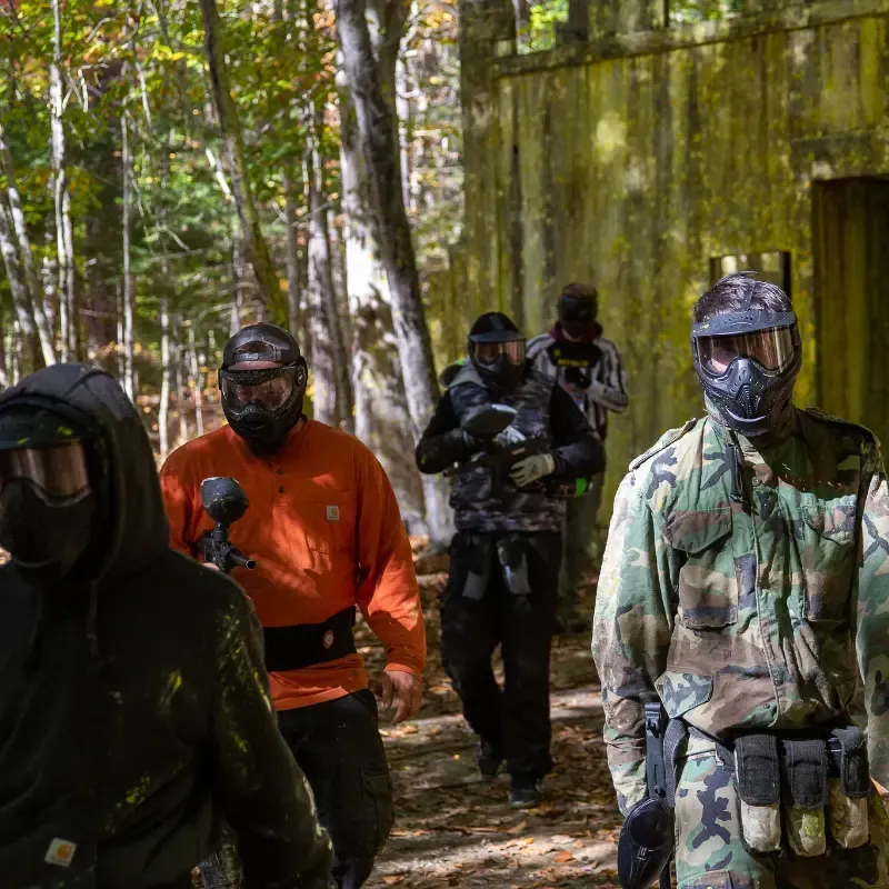 People in paintball gear walk near a bunker in a wooded area.