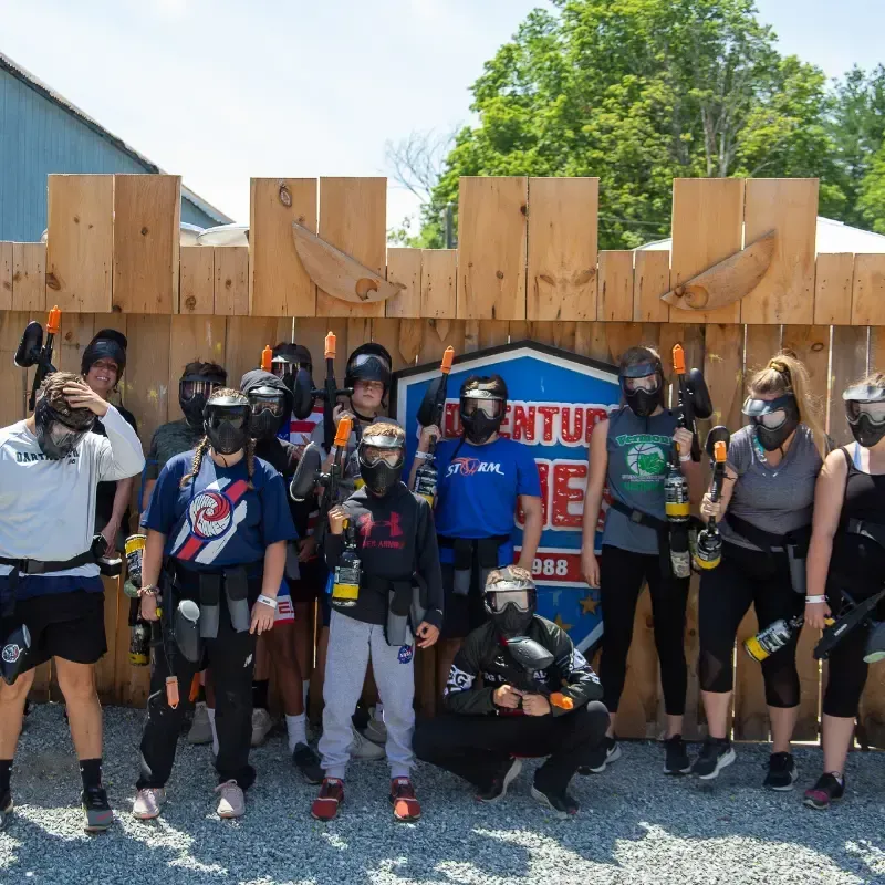 Group of people wearing paintball gear posing in front of a wooden structure.
