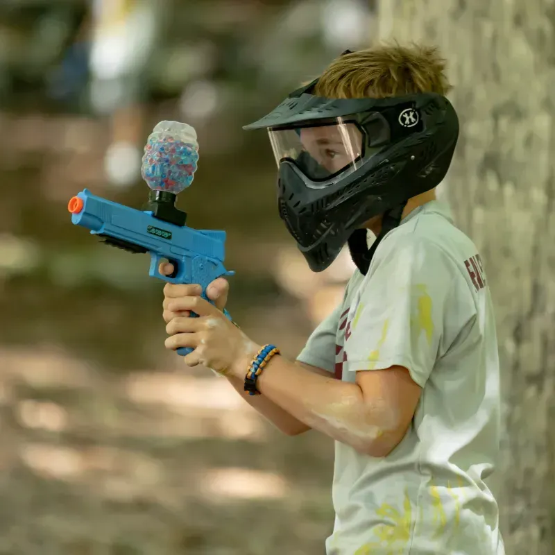 Boy in paintball mask aiming a blue paintball gun outdoors, covered in paint.