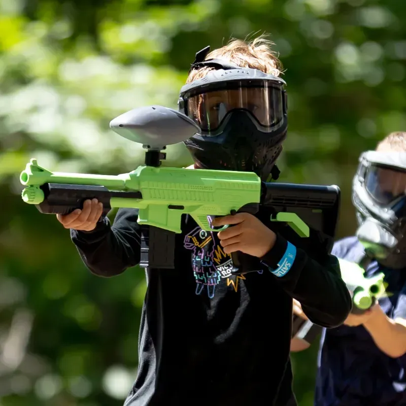 A child in black clothing and a paintball mask holding a neon green paintball gun outdoors.