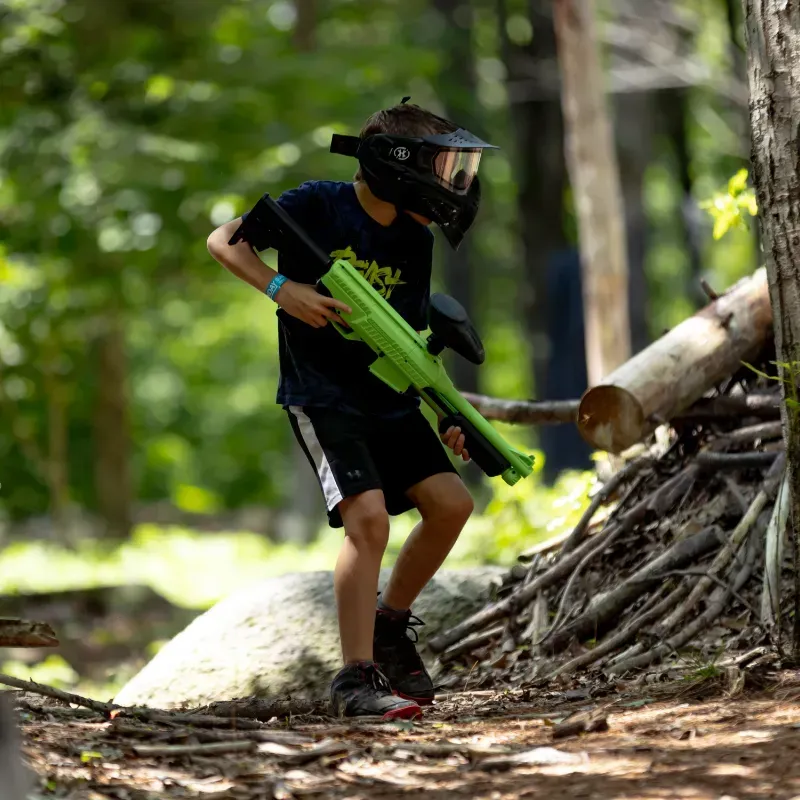 Young person in a helmet and holding a green paintball gun in a wooded area.