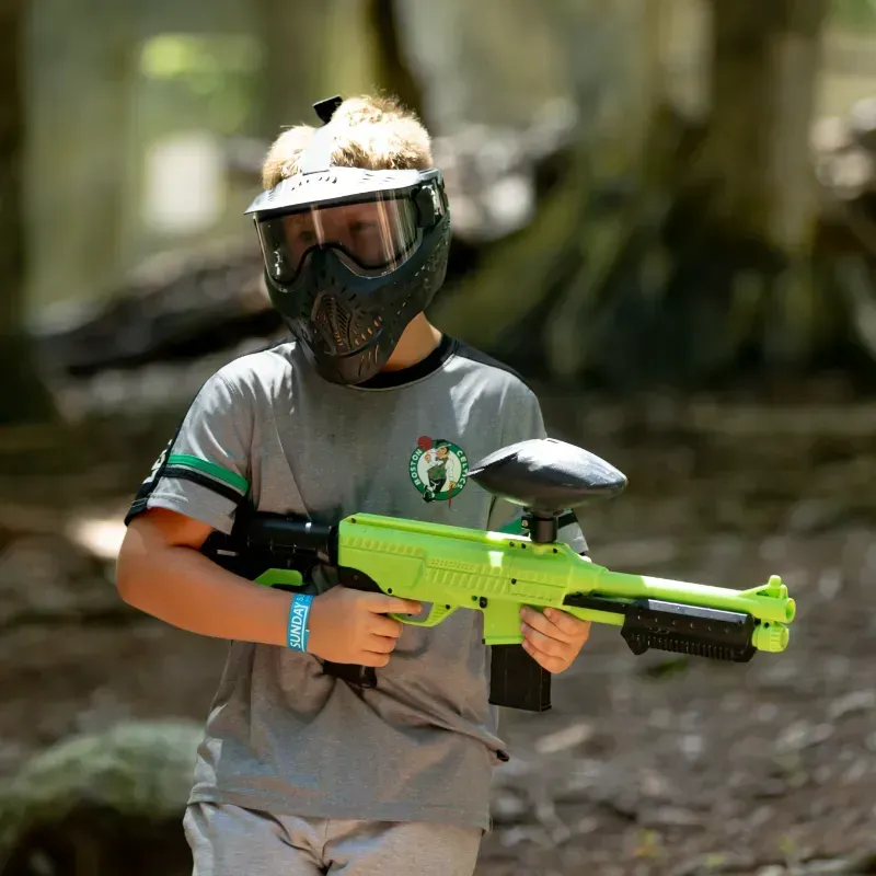 Boy in paintball mask and grey shirt holds a green paintball gun in a wooded area.