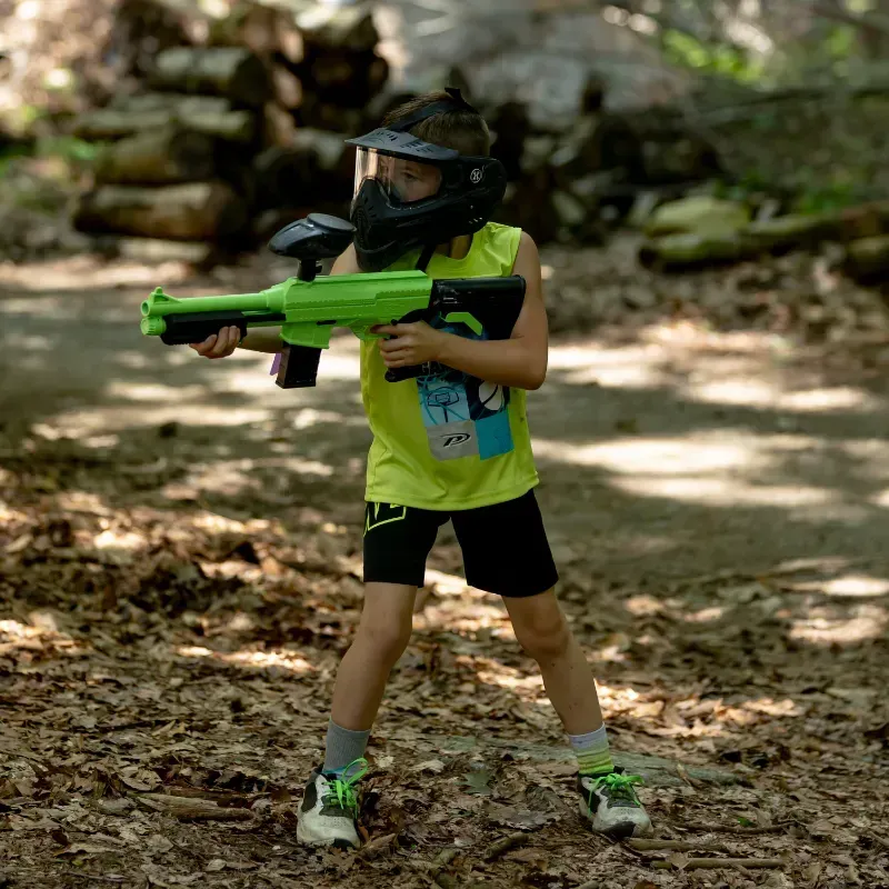 Boy in paintball gear holding a green gun, standing outdoors on a forest floor.