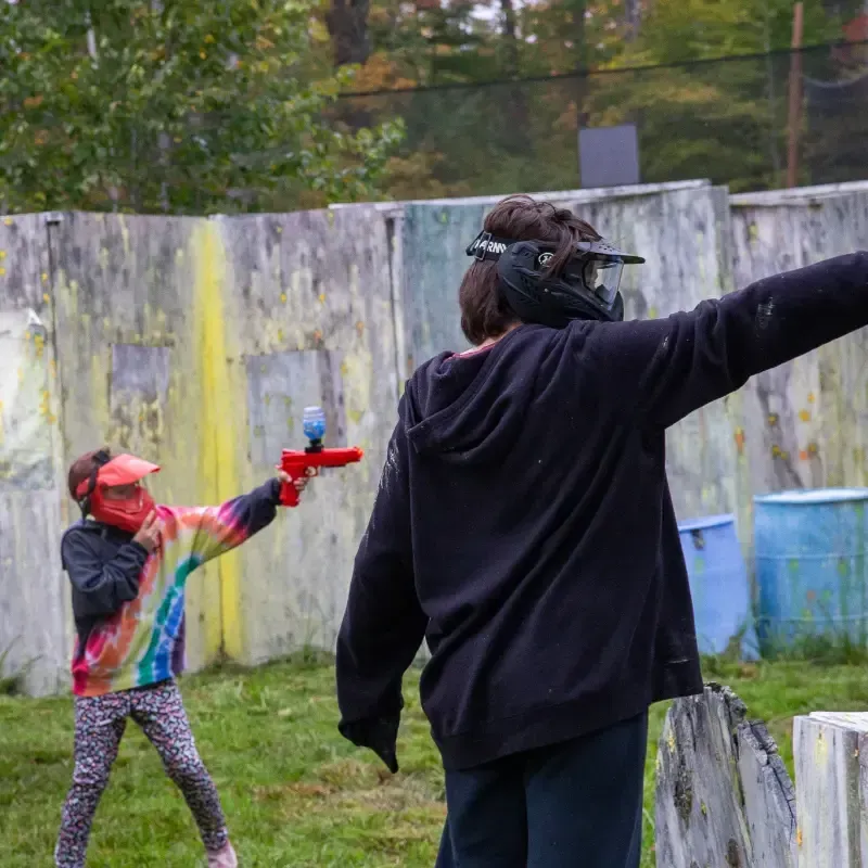 Two people playing paintball; one aims red gun at the other. They wear protective masks, in an outdoor course.
