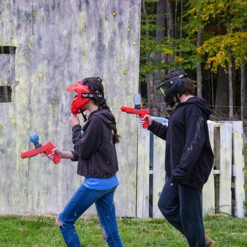 Two people in paintball gear play in a wooded area, holding red guns.