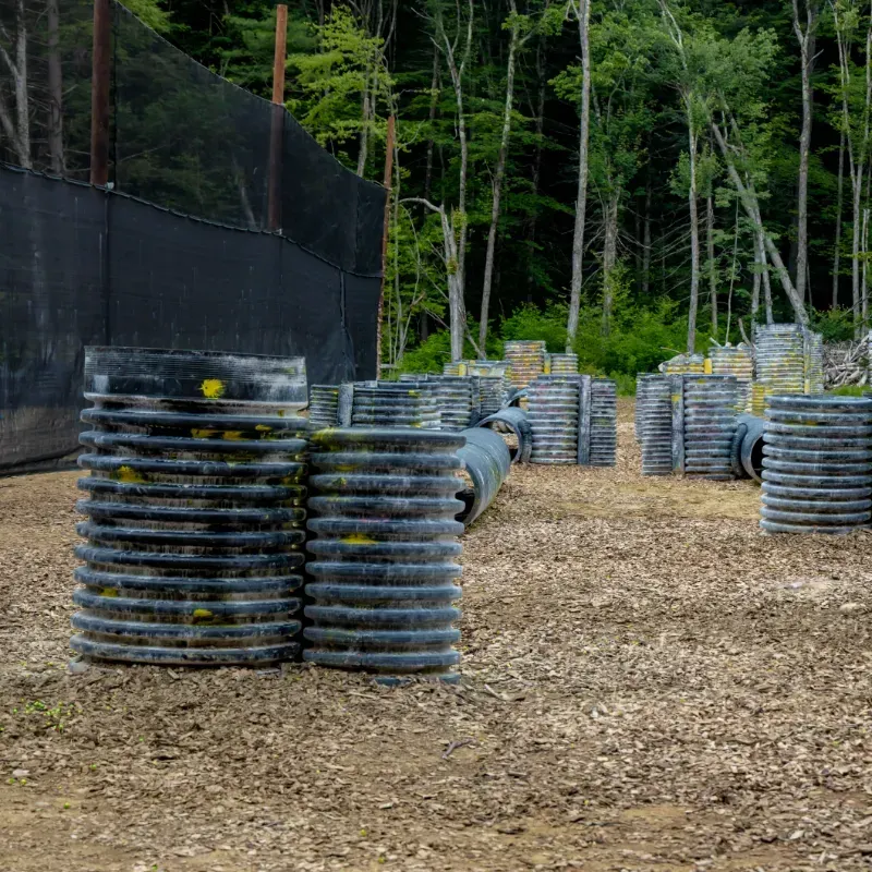 Paintball course with black barrels and protective netting in a forest setting.