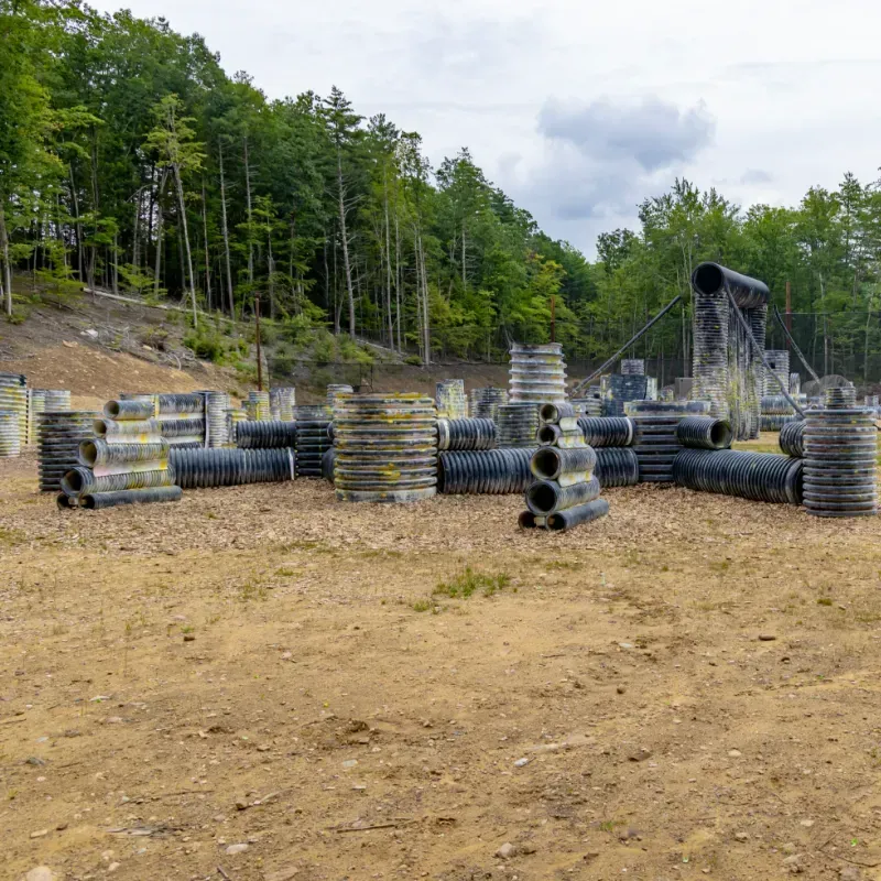 Paintball field with tire and tube bunkers, set against a wooded hillside under a cloudy sky.