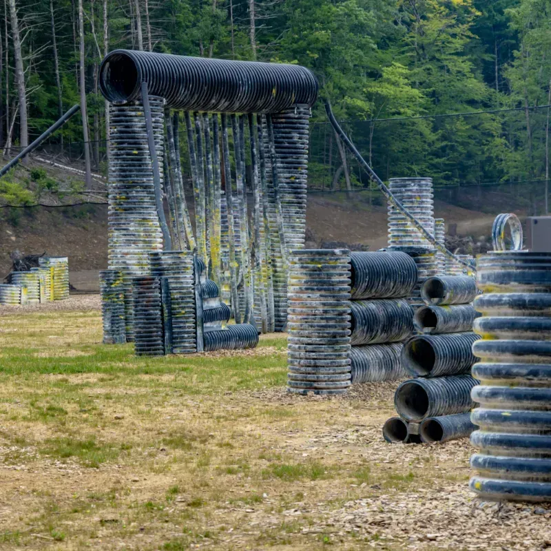 Paintball field with black and gray tube obstacles in a grassy area, with a forest background.