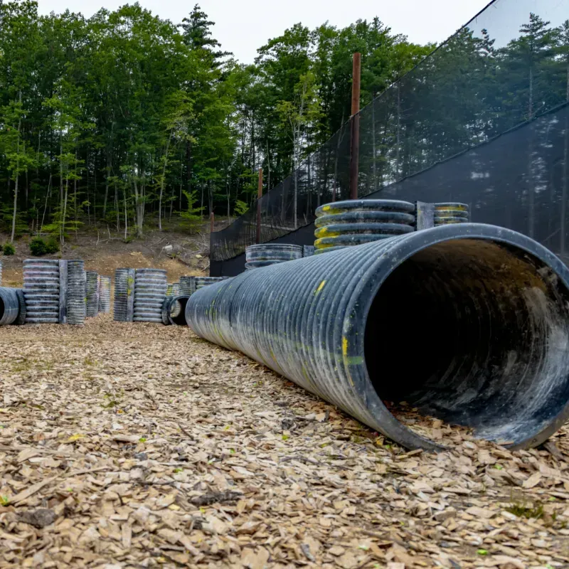 Paintball course with large black culvert, tires, and wooden obstacles. Green trees in background.